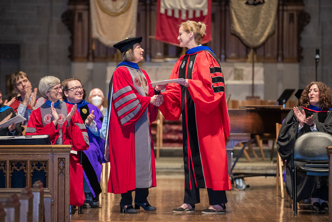 A woman in academic robes and a hat shakes hands with a tall woman in academic robes with blonde hair